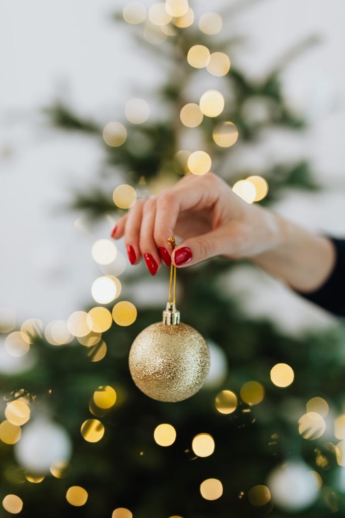 12 Christmas Nail Colors for 2026 (With Exact Polish Names) Classic red Christmas nails holding a gold glitter ornament in front of a Christmas tree