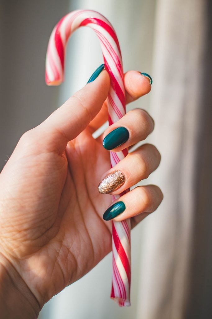 12 Christmas Nail Colors for 2026 (With Exact Polish Names) Dark green Christmas nails with a gold glitter accent nail holding a candy cane